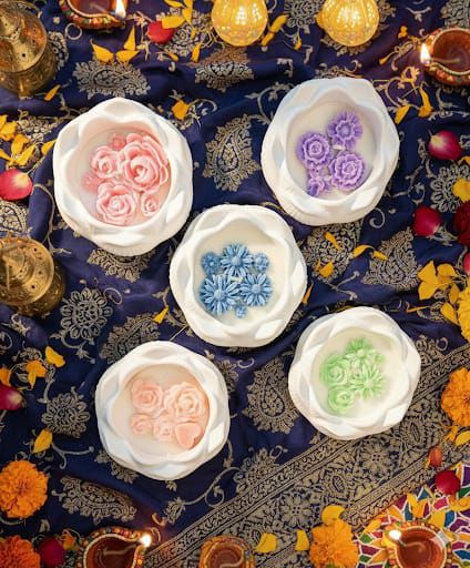 Pastel floral candles in ceramic containers arranged on festive fabric with diyas and flower petals